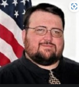 Head shot of Joseph "Joe" Terrell in a blue dress shirt standing in front of a white backdrop and a U.S. Flag.